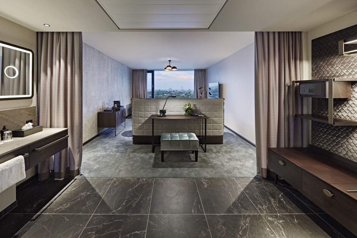 Habitación de hotel moderna con sofá, una mesita, un banco, televisor, minibar y vistas a la ciudad.