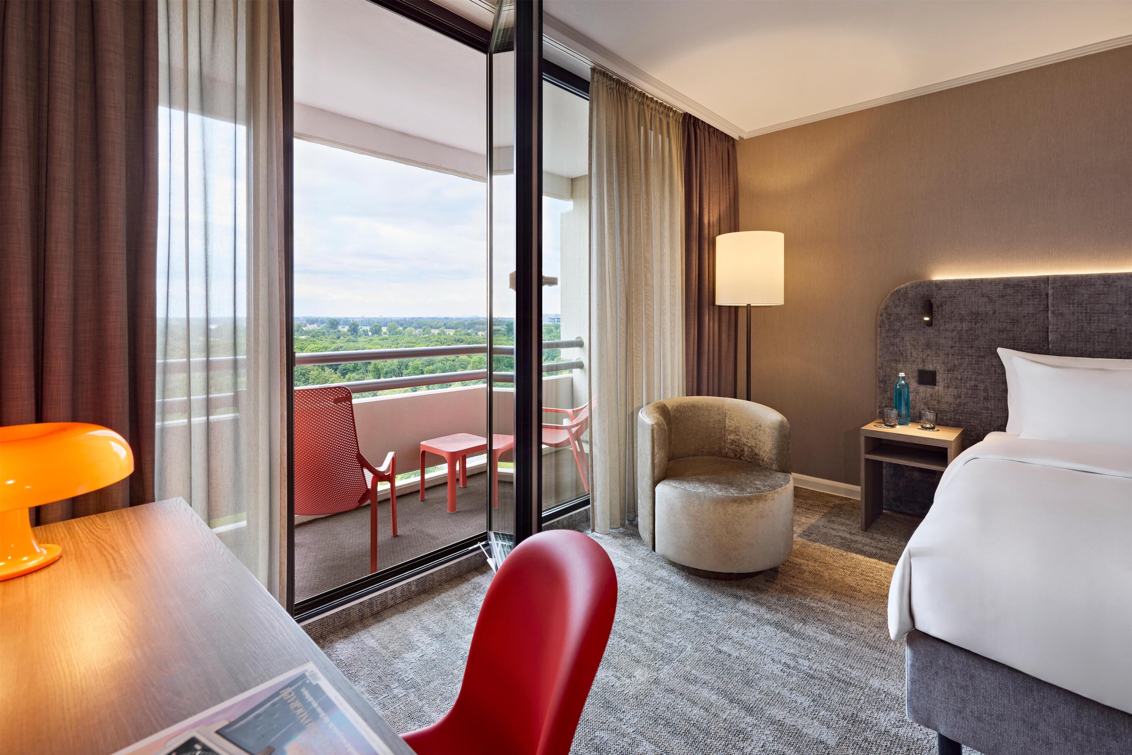Habitación de hotel con balcón, cama, silla, escritorio y lámpara.