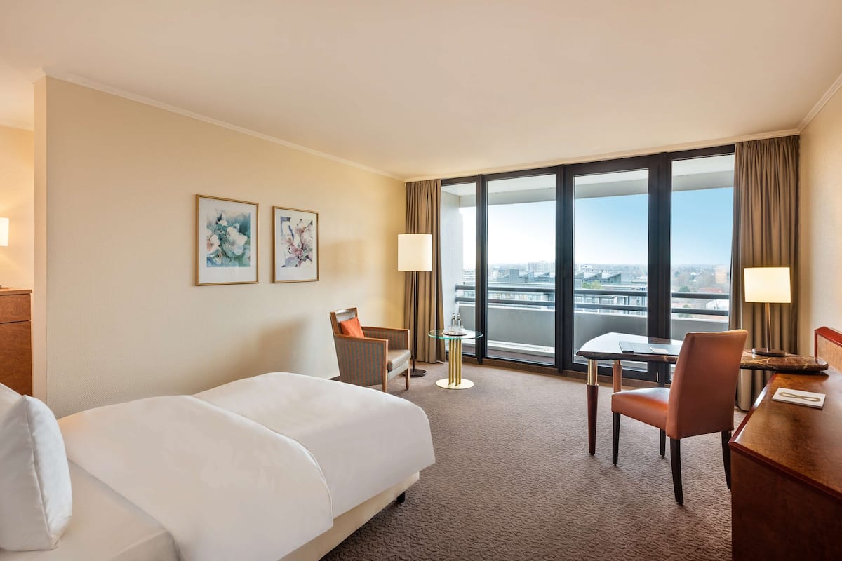 A hotel room with a large bed, a desk, two chairs, a small table, and a view of the city through large windows.