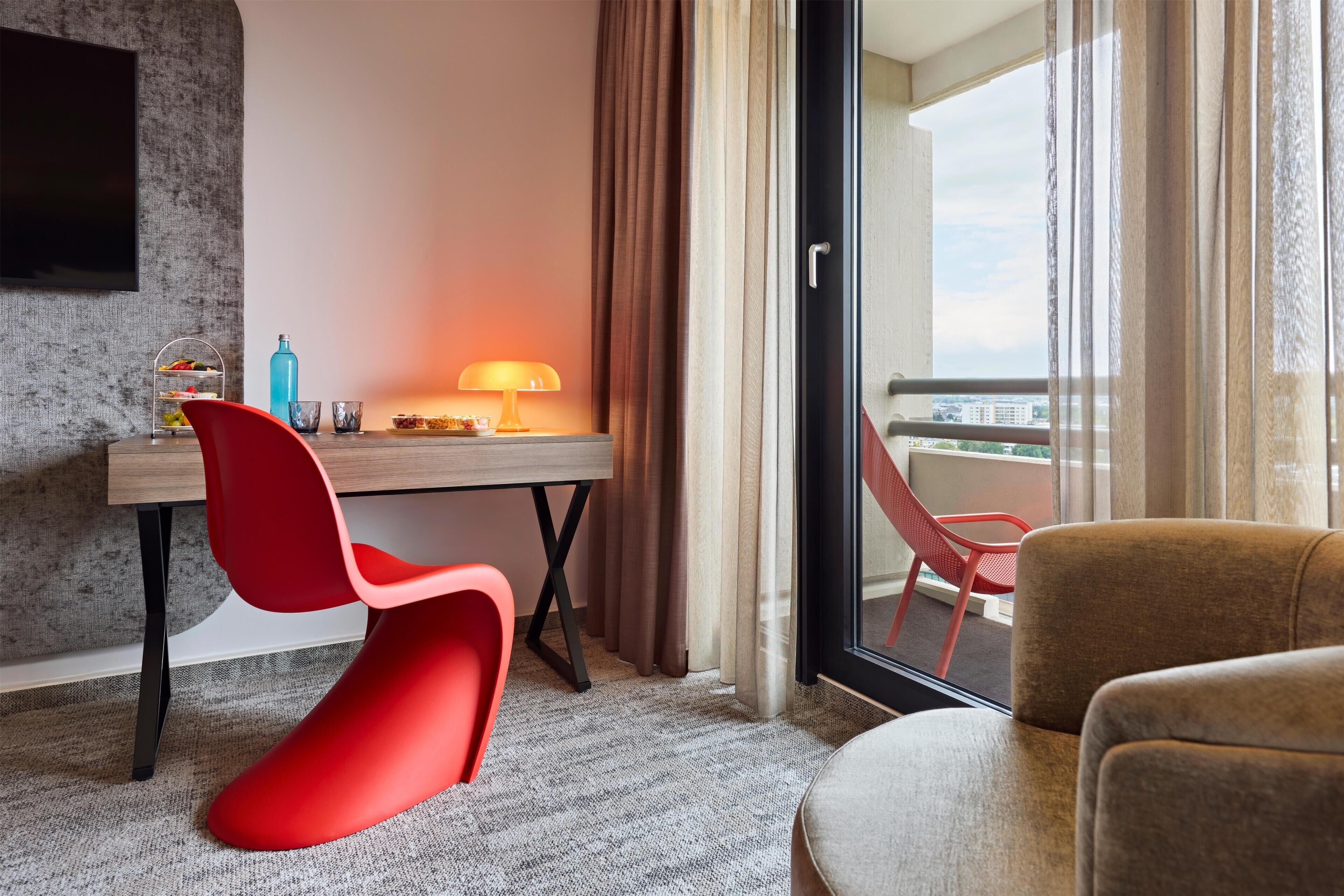 Habitación de hotel con escritorio, una silla roja, sofá, balcón con vistas y televisor montado en la pared.
