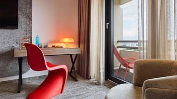 Habitación de hotel con escritorio, una silla roja, sofá, balcón con vistas y televisor montado en la pared.