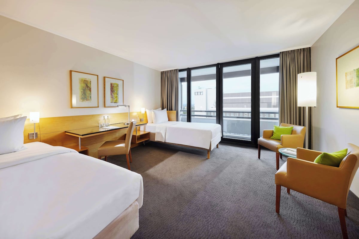 A hotel room with two beds, a desk, a chair, a small table, and a view of the city through large windows.