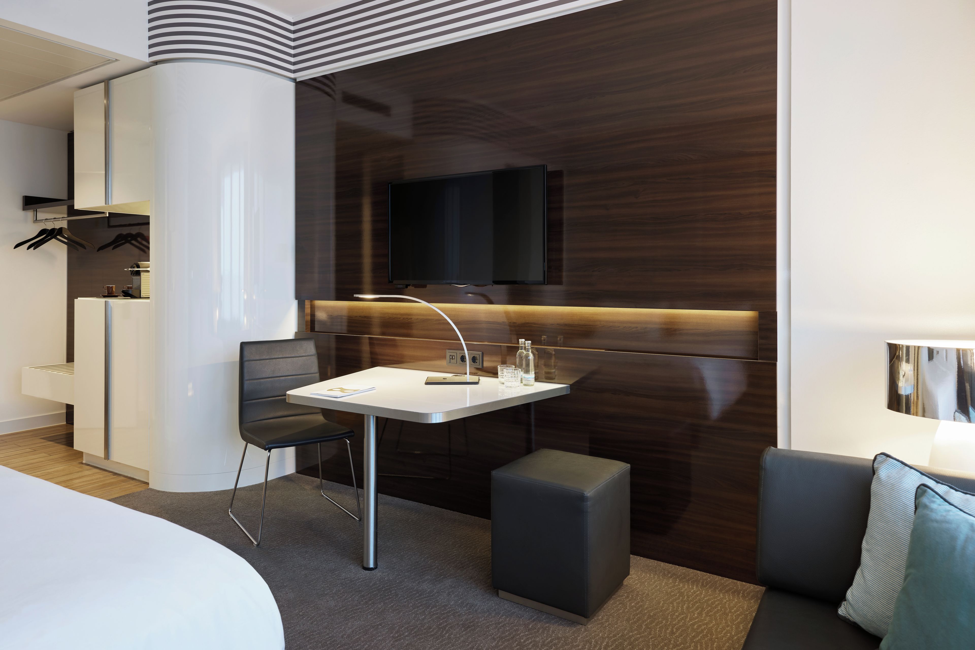 Habitación de hotel moderna con una cama blanca, un escritorio con una silla, un pequeño otoman, un televisor de pantalla plana montado en una pared de madera oscura y una lámpara a la derecha.