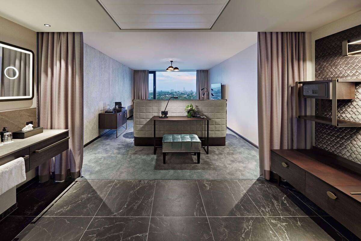 A modern hotel room with a sofa, a small table, a bench, a desk, a mirror, a TV, and a city view through the window.