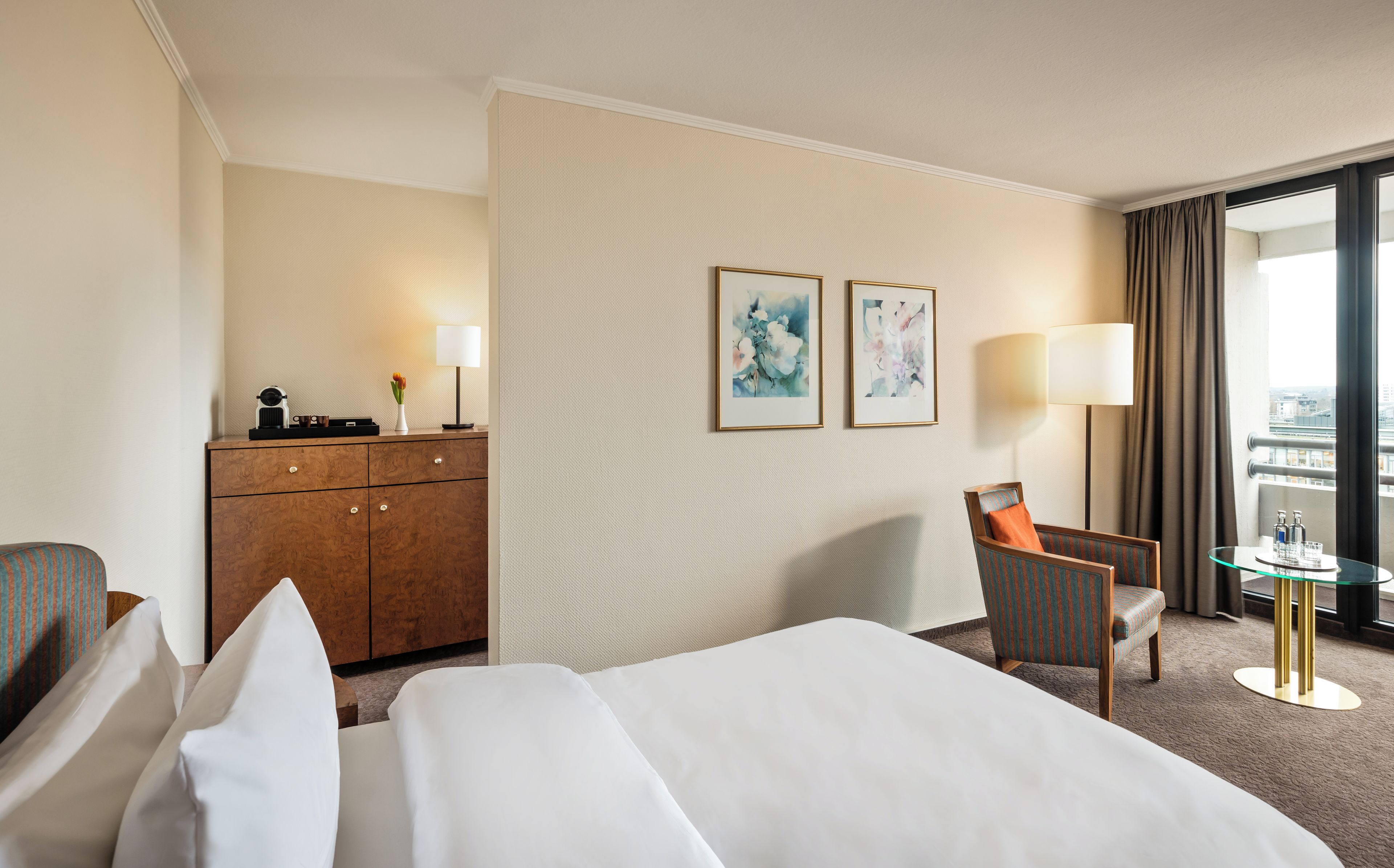 Una habitación de hotel con una cama, un escritorio con lámpara, una silla y dos cuadros en la pared.