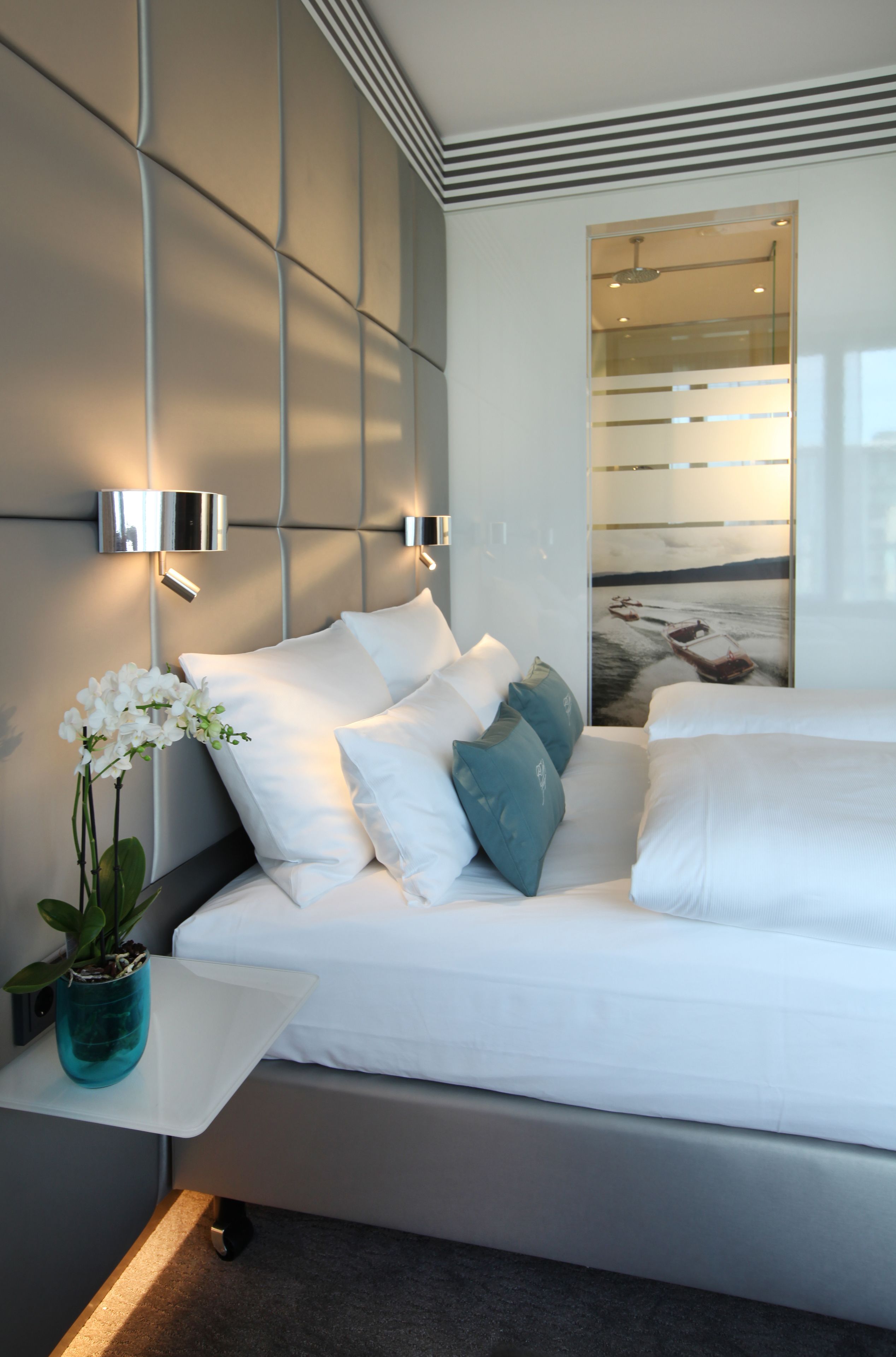 Une chambre moderne avec un lit, du linge de lit blanc, une orchidée en pot et un luminaire fixé au mur.