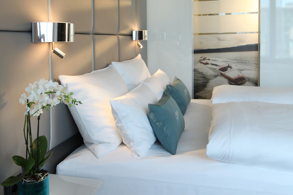 A modern bedroom with a bed, white bedding, a potted orchid, and a wall-mounted light fixture.