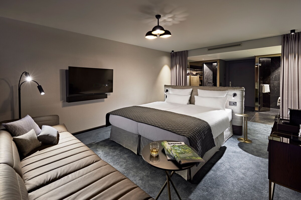 A modern hotel room with a large bed, a sofa, a TV, and a small table with a magazine.