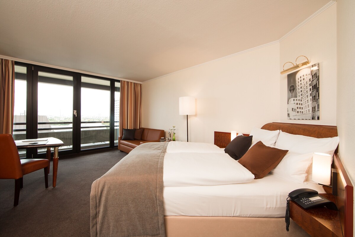 A hotel room with a large bed, a desk, a chair, and a view of the city through large windows.