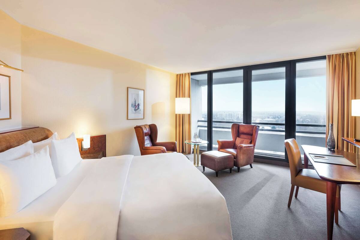A hotel room with a large bed, two armchairs, a small table, and a view of the city through large windows.
