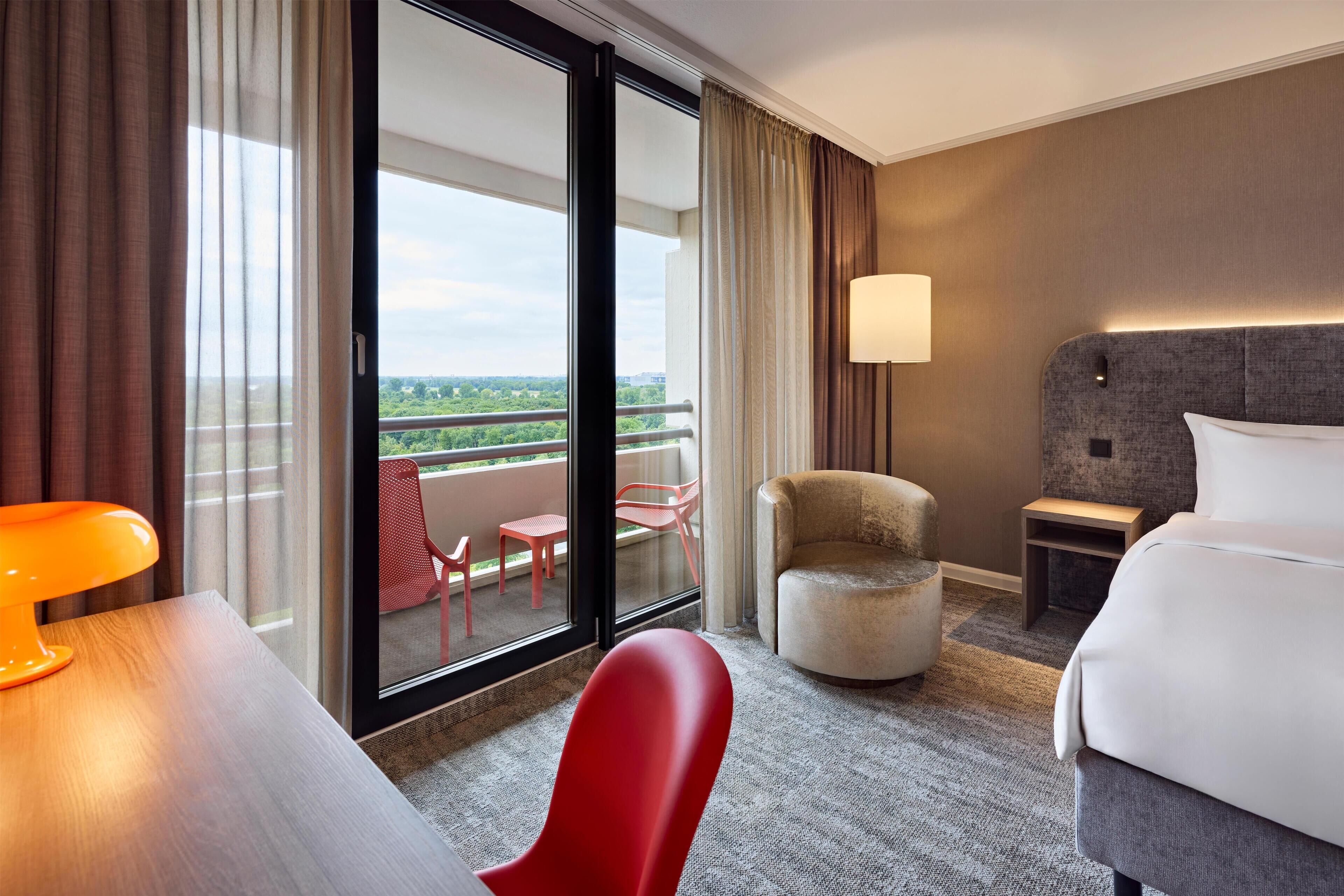A hotel room with a large window, a bed, a desk, a chair, a lamp, and a view of greenery outside.