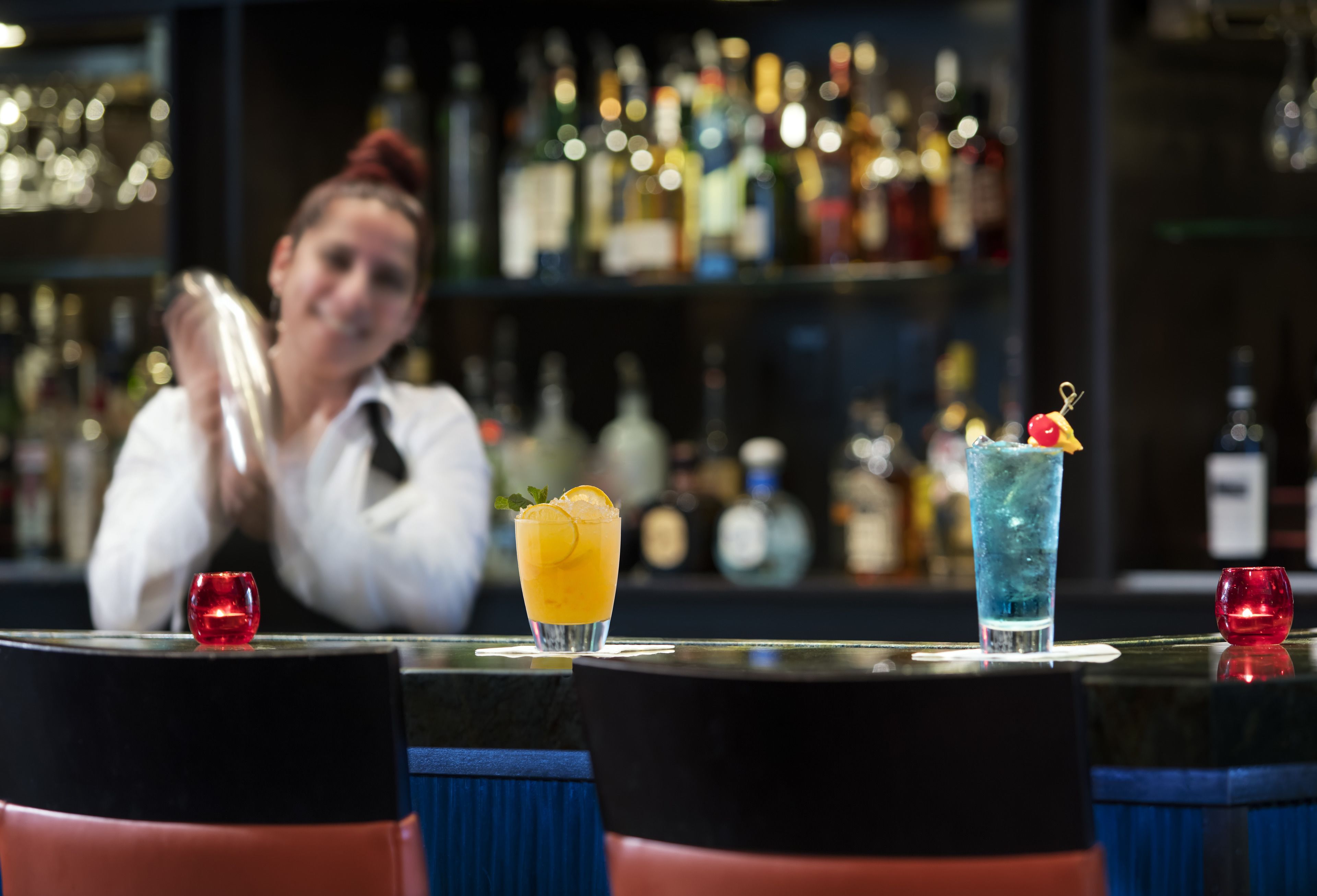 A bar with two drinks on the counter, a bartender in the background, and shelves of bottles behind.