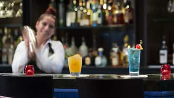 A bar with two drinks on the counter, a bartender in the background, and shelves of bottles behind.