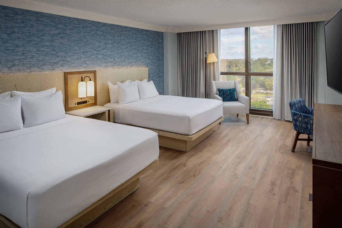 A hotel room with two beds, a desk, a chair, a TV, and a large window with curtains.