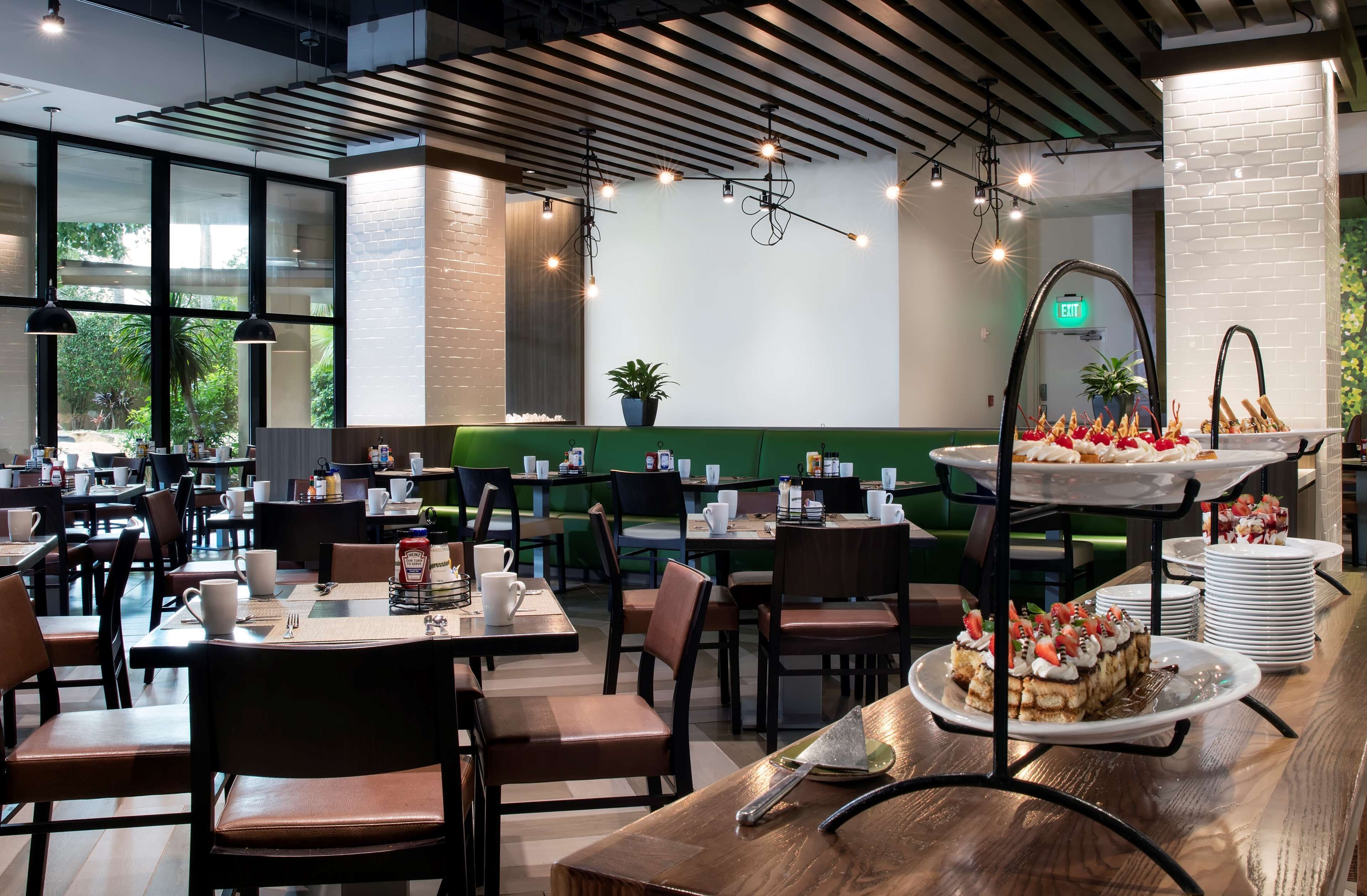 A modern restaurant with high ceilings, exposed beams, and large windows. Tables are set with white plates, silverware, and cups. A dessert station with a cake and strawberries is visible.