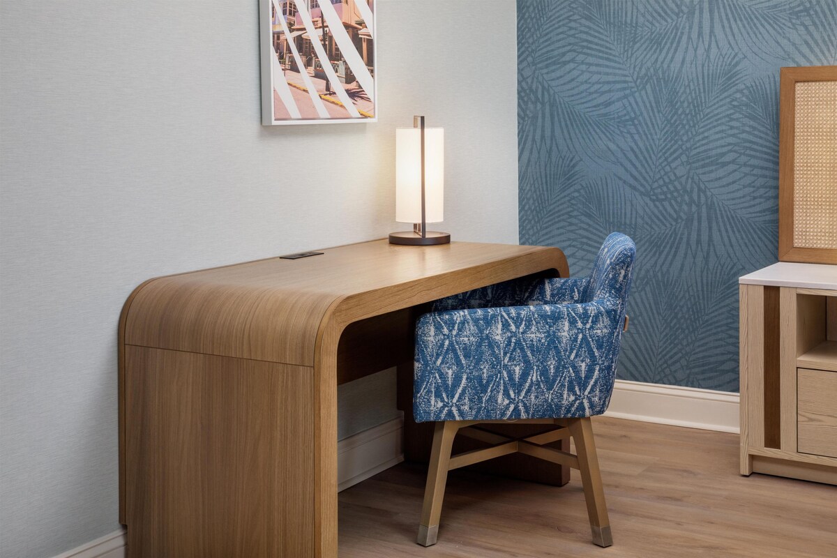 A wooden desk with a lamp, a blue patterned chair, and a small cabinet.