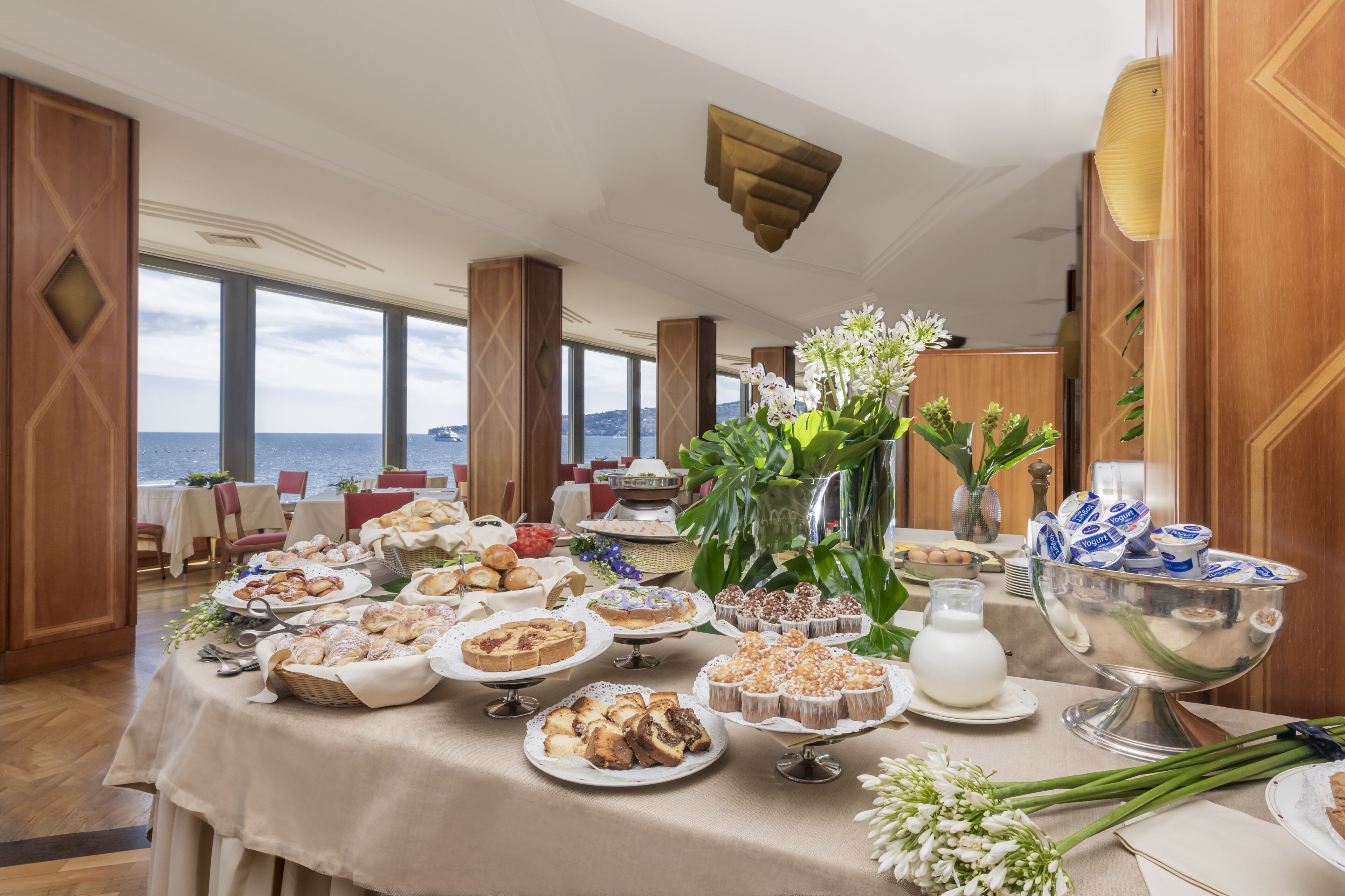 Een buffettafel met diverse gebakjes en desserts, een bloemenvaas en uitzicht op zee door grote ramen.