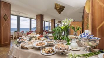 A buffet table with various pastries and desserts, a vase with flowers, and a view of the sea through large windows.