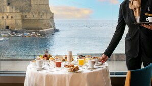 A breakfast table with various food items and beverages, overlooking a coastal view with a historic building. - Royal Continental Hotel Naples (Naples)