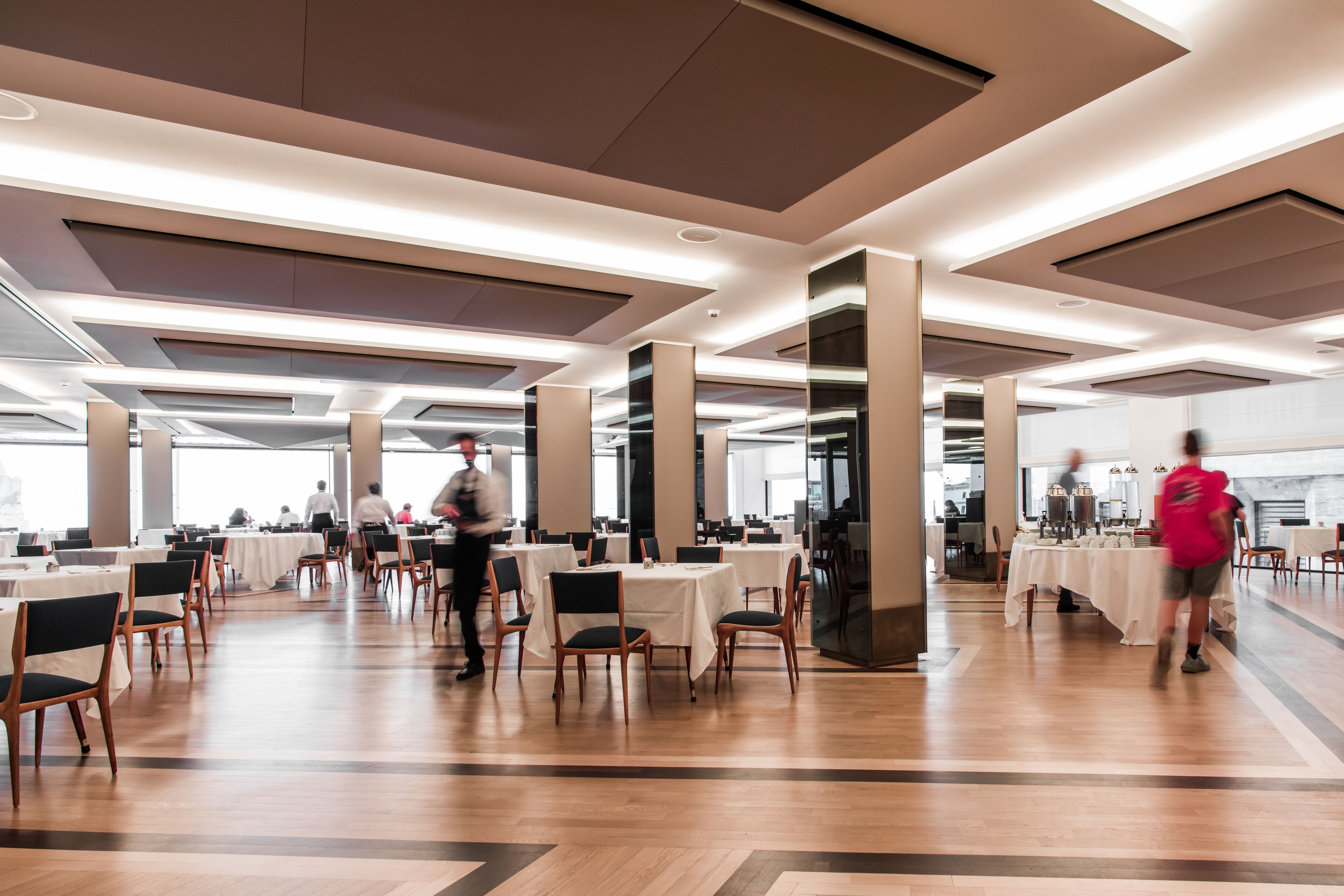 A spacious dining area with multiple tables and chairs, high ceilings with recessed lighting, and a modern architectural design.