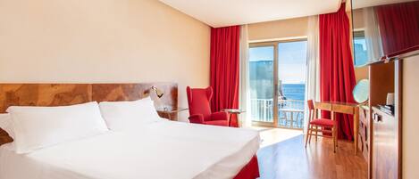 A hotel room with a large bed, a desk with a chair, and a view of the sea through the balcony.