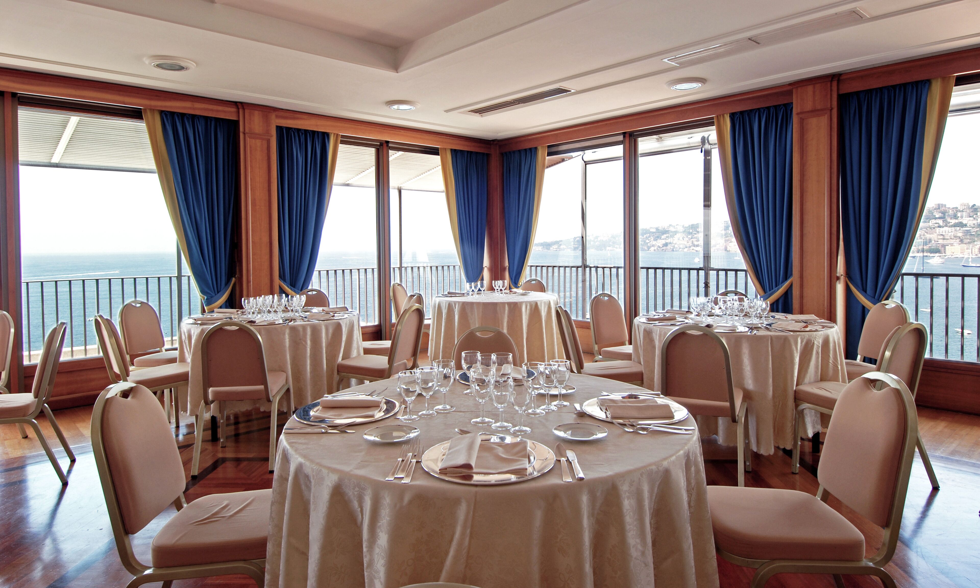 A restaurant with round tables set for dining, large windows with blue curtains, and a view of the sea.