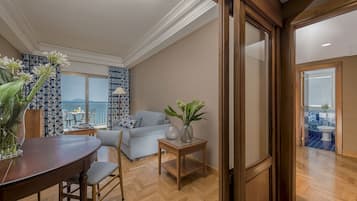 Een moderne woonkamer met een houten eettafel, een bank en een kleine bijzettafel met een vaas. Een groot raam biedt uitzicht op zee.