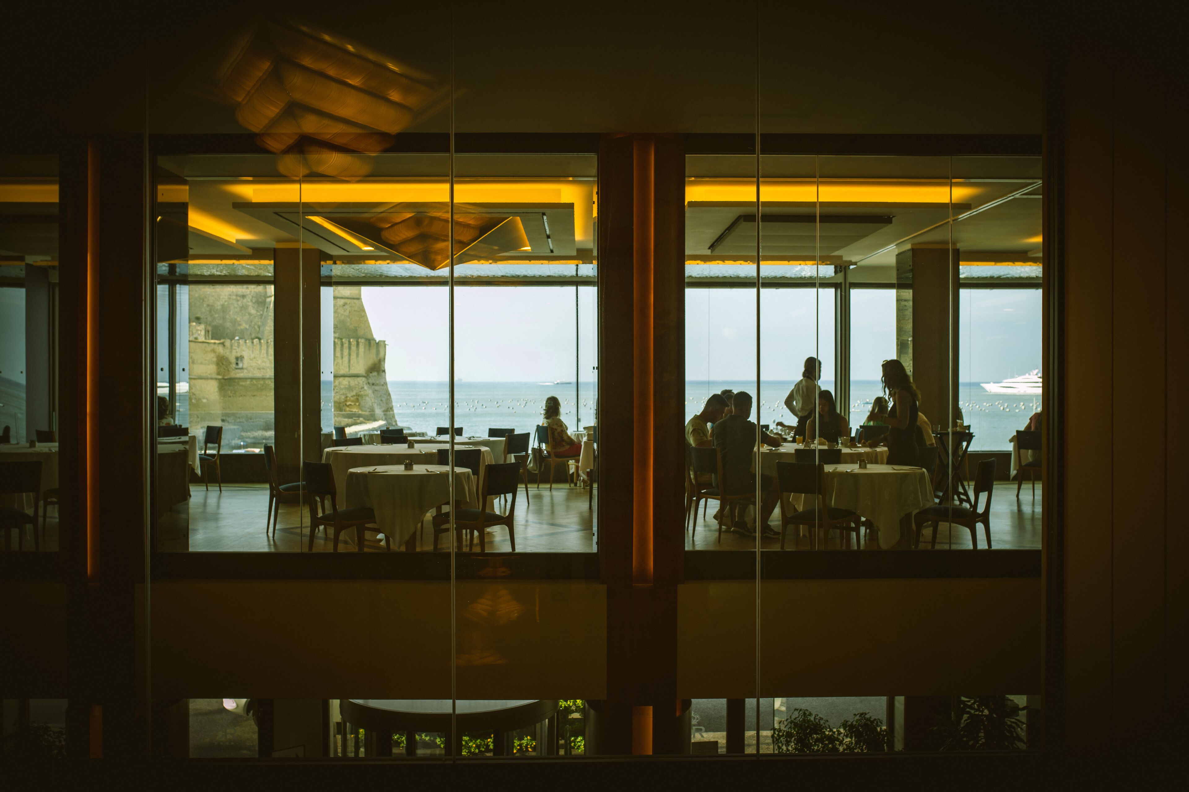 A restaurant with large windows overlooking a coastal view, featuring tables and chairs arranged for dining.