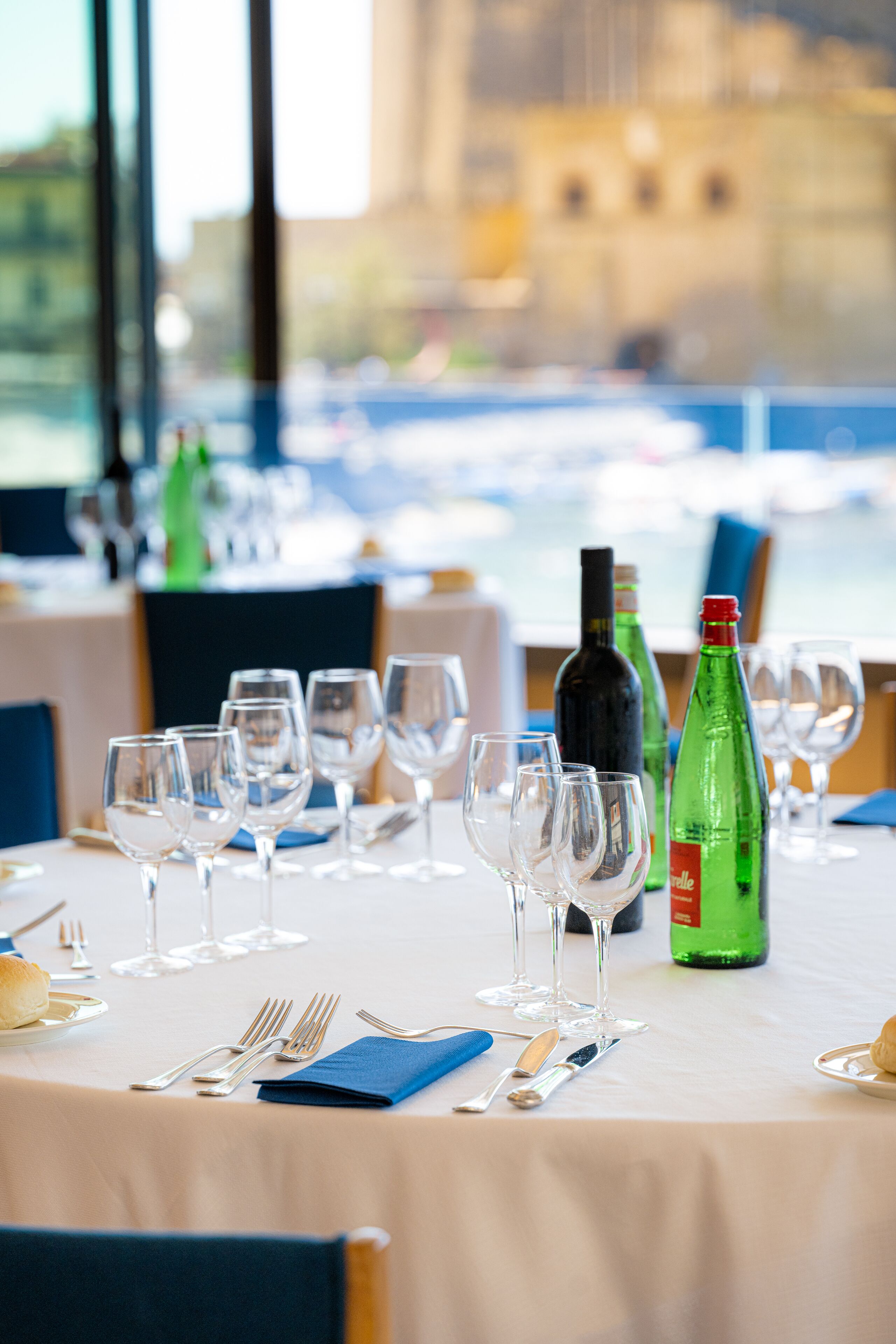 A dining table set with wine glasses, bottles, and cutlery, overlooking a marina.