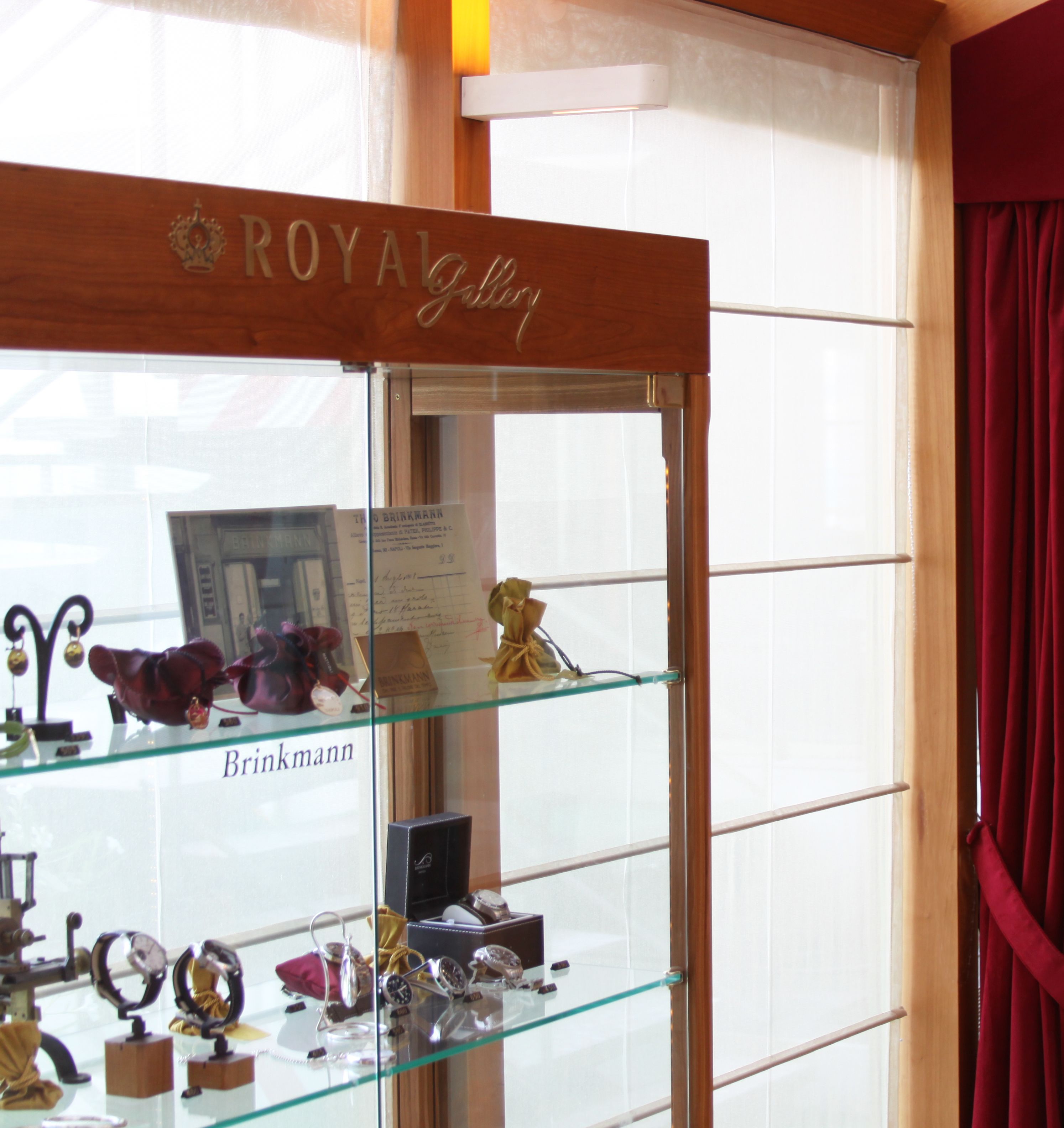 Una vitrina de vidrio con diversos artículos, entre ellos relojes y joyería, en una habitación con techo de madera y una ventana con cortinas rojas.
