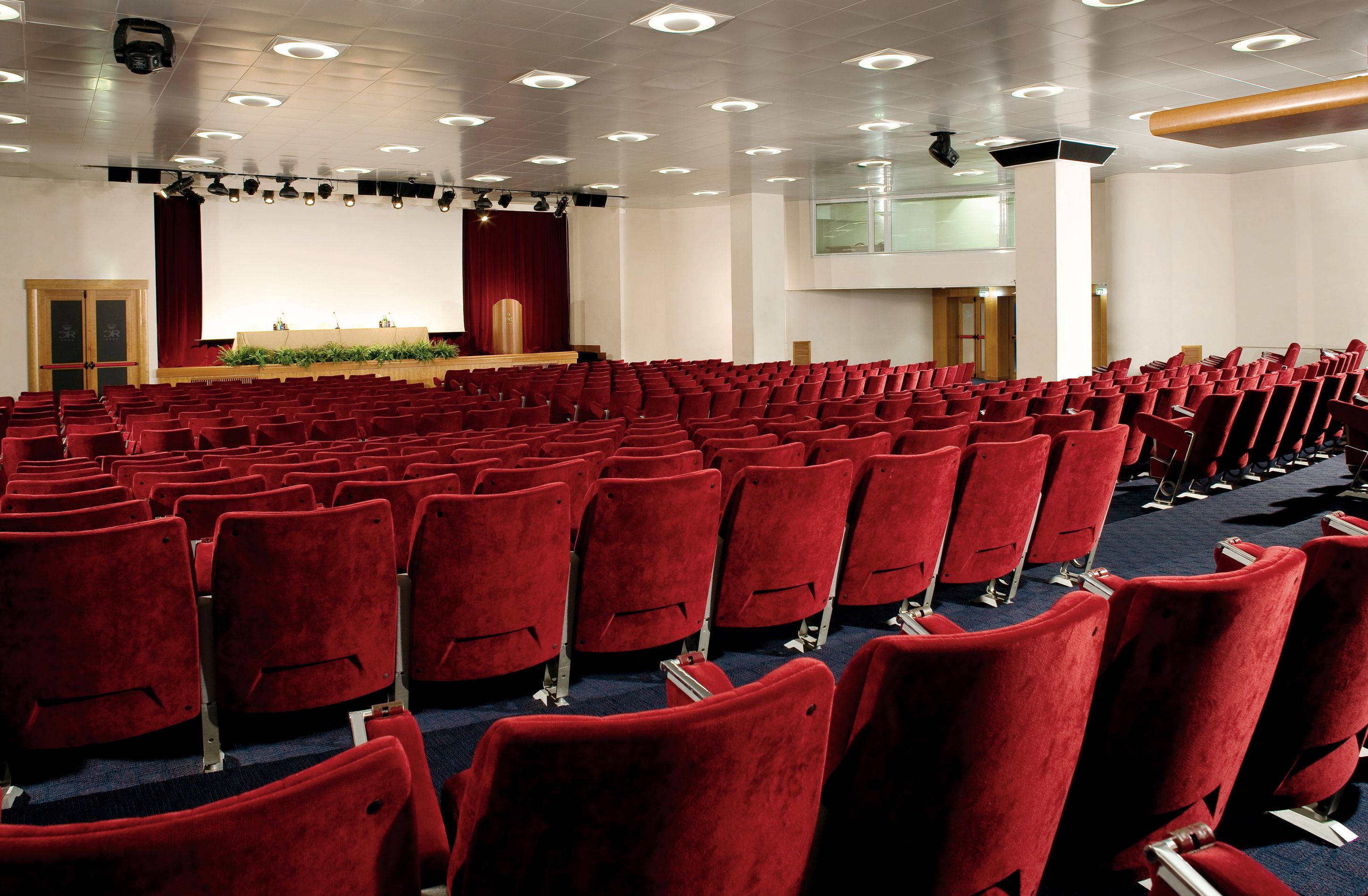 A large auditorium with rows of red chairs, a stage with a red curtain, and a projector screen.