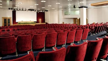 A large auditorium with rows of red chairs, a stage with a red curtain, and a projector screen.