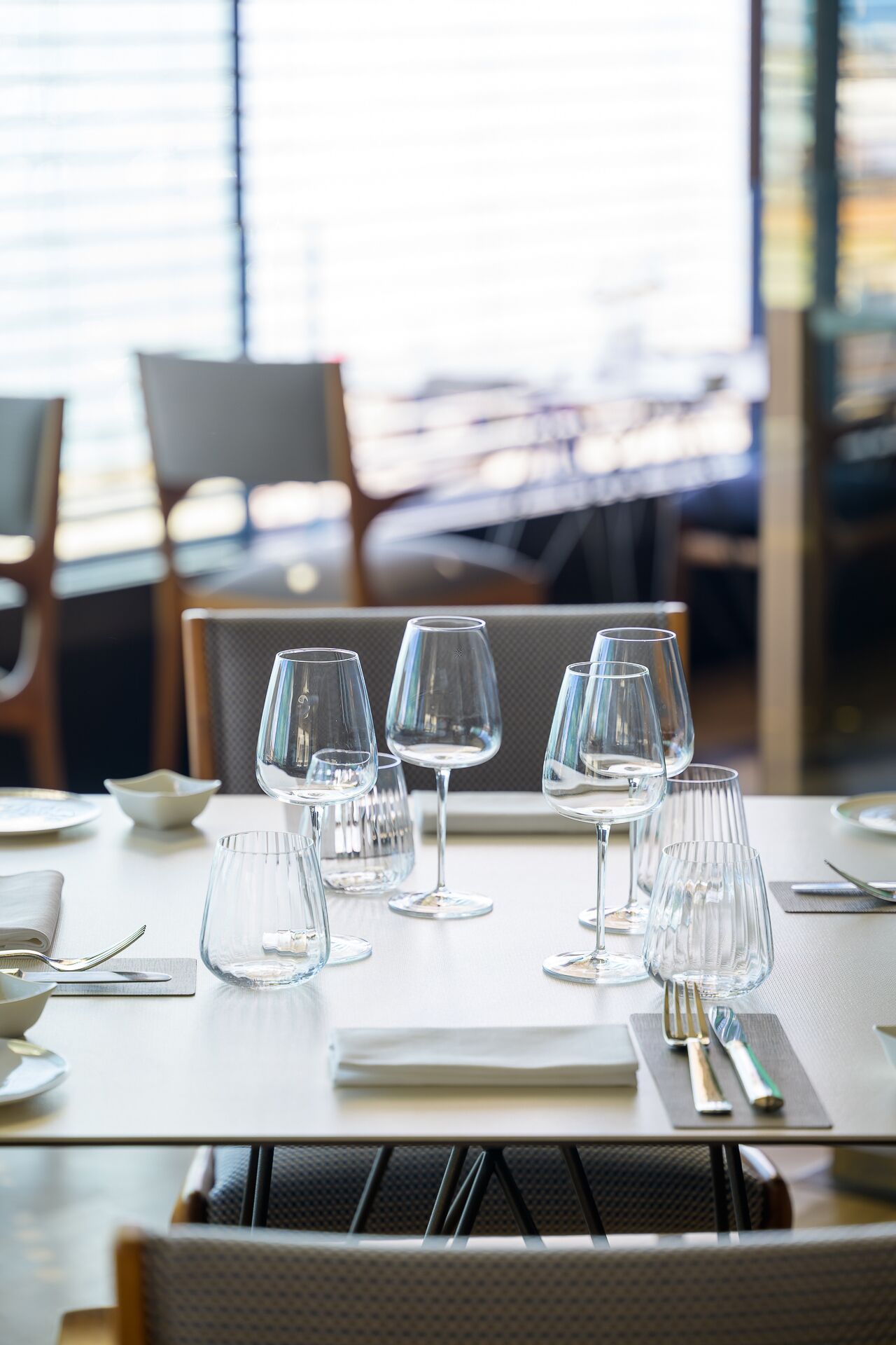 A dining table set with multiple wine glasses and cutlery, with chairs and a window in the background.