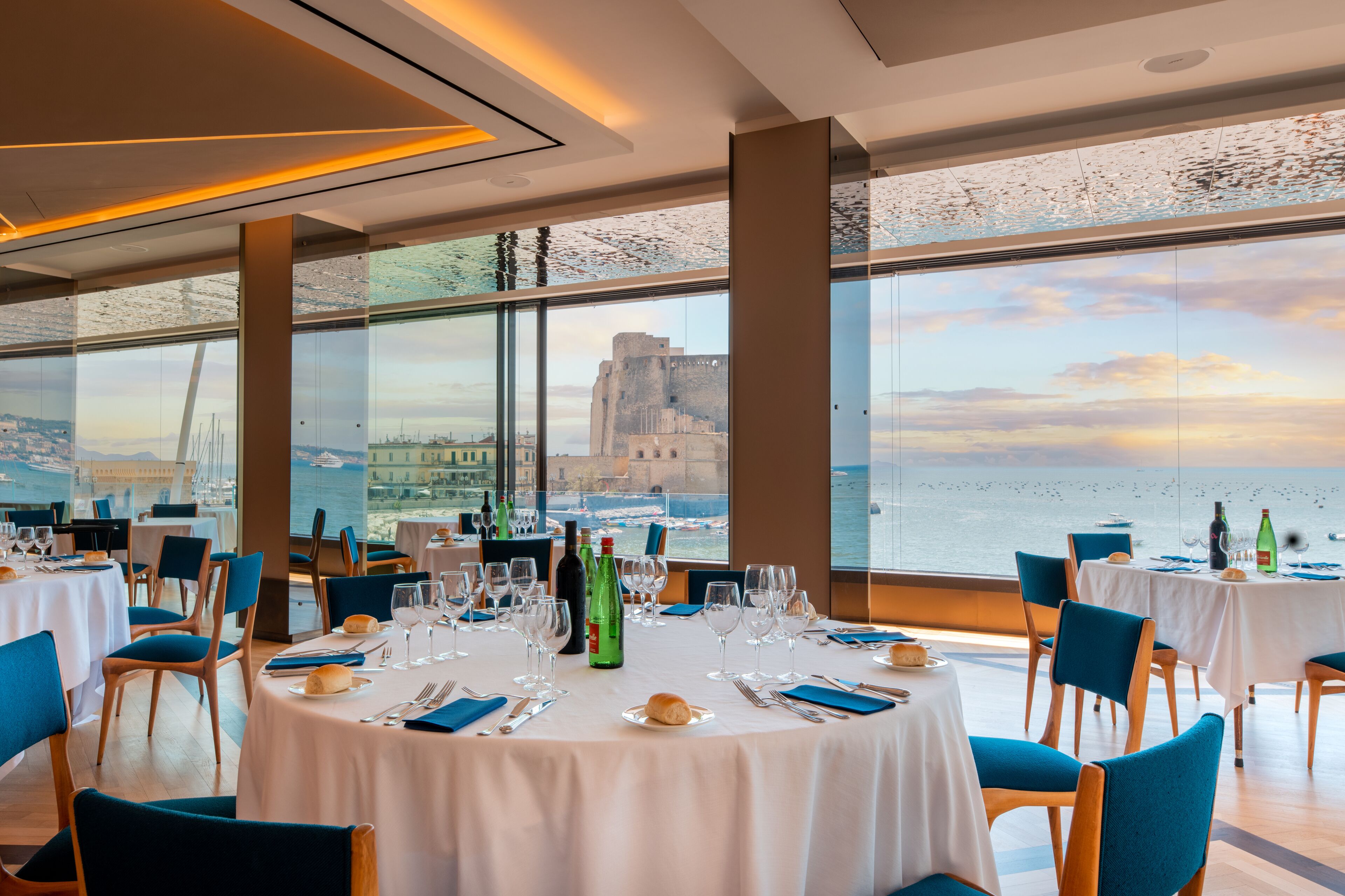 Un restaurant amb taules rodones preparades per a dinar, grans finestres que ofereixen vista al mar i un edifici distant.