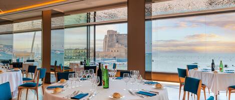 Un restaurant avec des tables rondes dressées pour le dîner, de grandes fenêtres offrant une vue sur la mer et un bâtiment situé au loin.