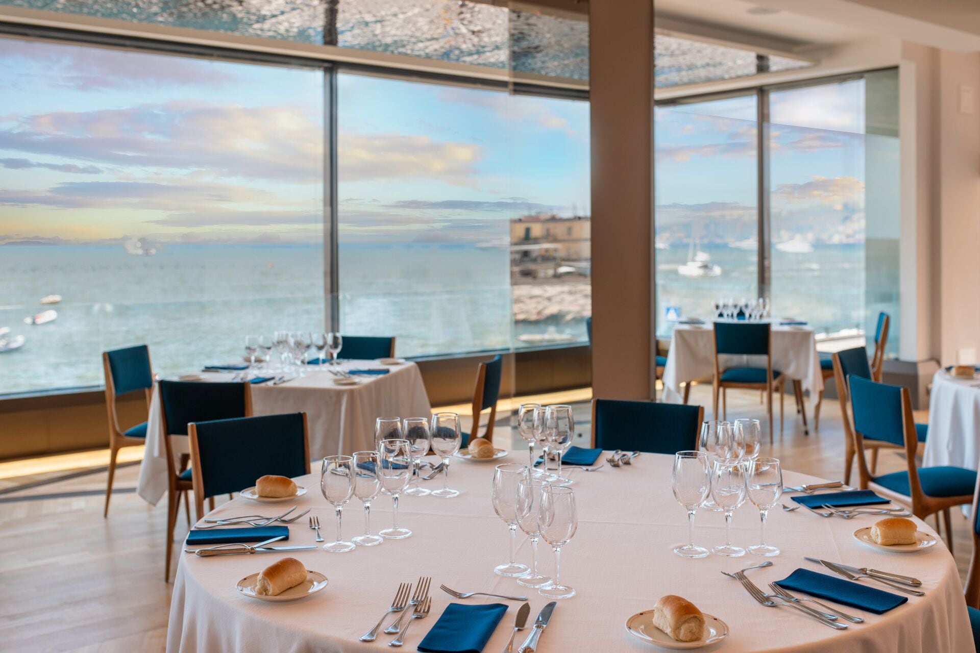 A dining area with tables set for a meal, overlooking a body of water with boats.