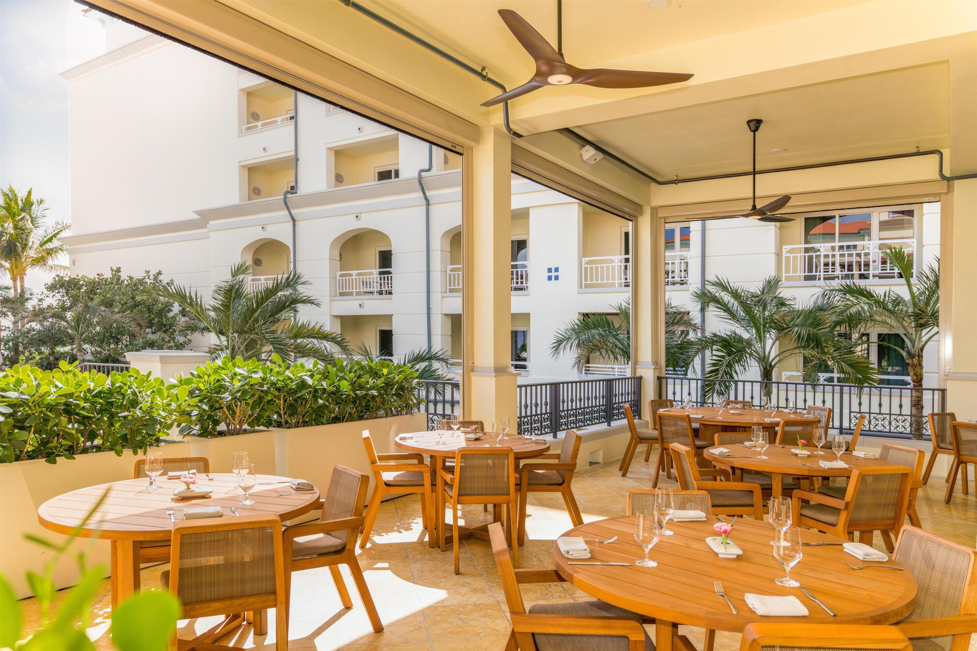 A restaurant with round tables and chairs, large windows, and a view of a building with balconies.