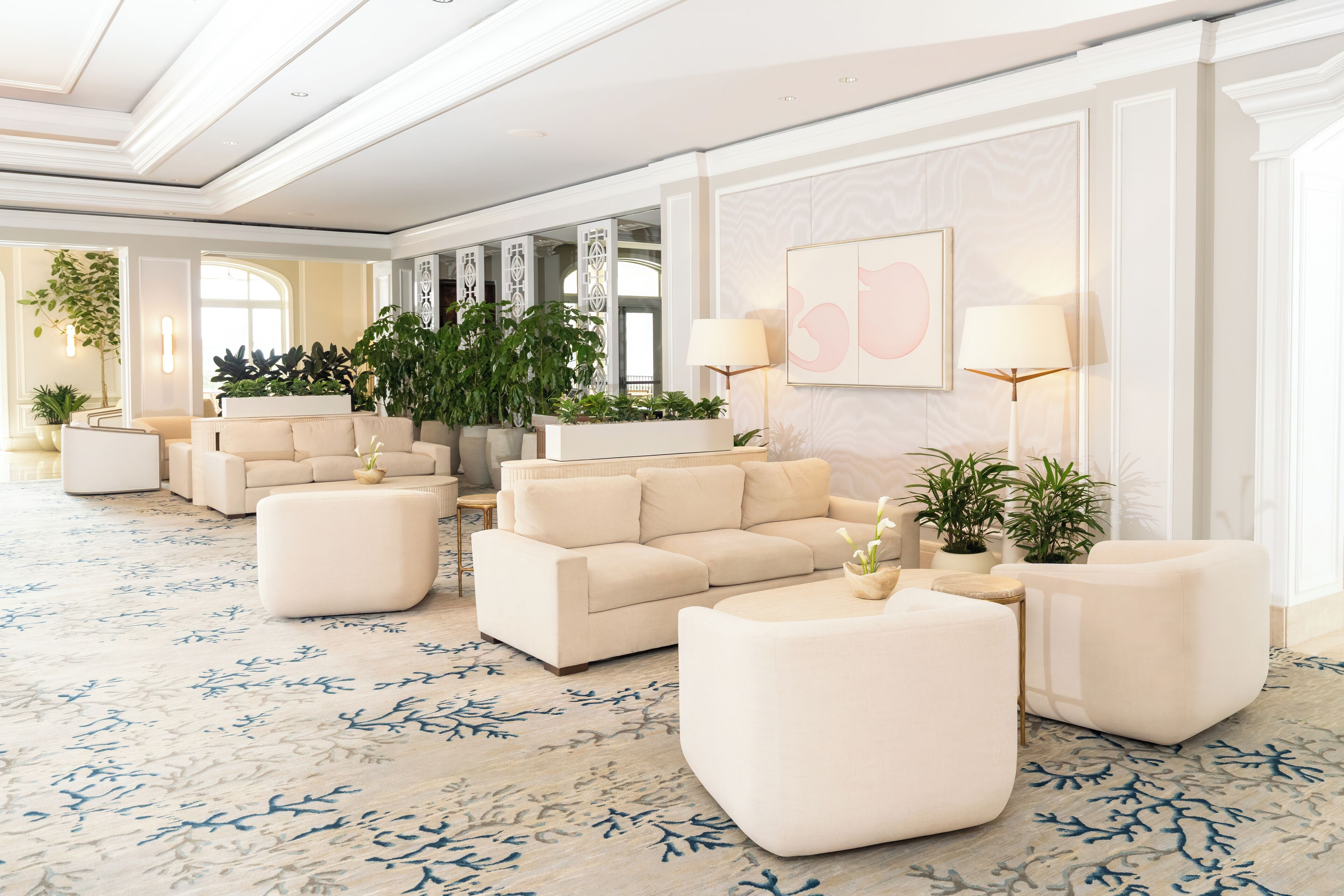 A spacious lobby with a modern design, featuring a large seating area with sofas and ottomans, a patterned carpet, and several potted plants.