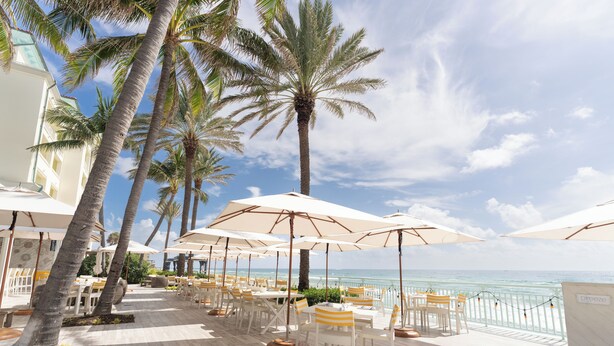 A seaside patio with white tables and chairs, palm trees, and umbrellas. - Eau Resort & Spa (Manalapan)