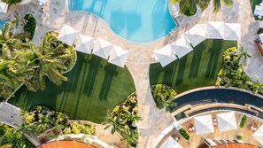 Luftaufnahme eines Resorts mit einem zentral gelegenen Schwimmbad, umgeben von Palmen und weißen Sonnenschirmen.