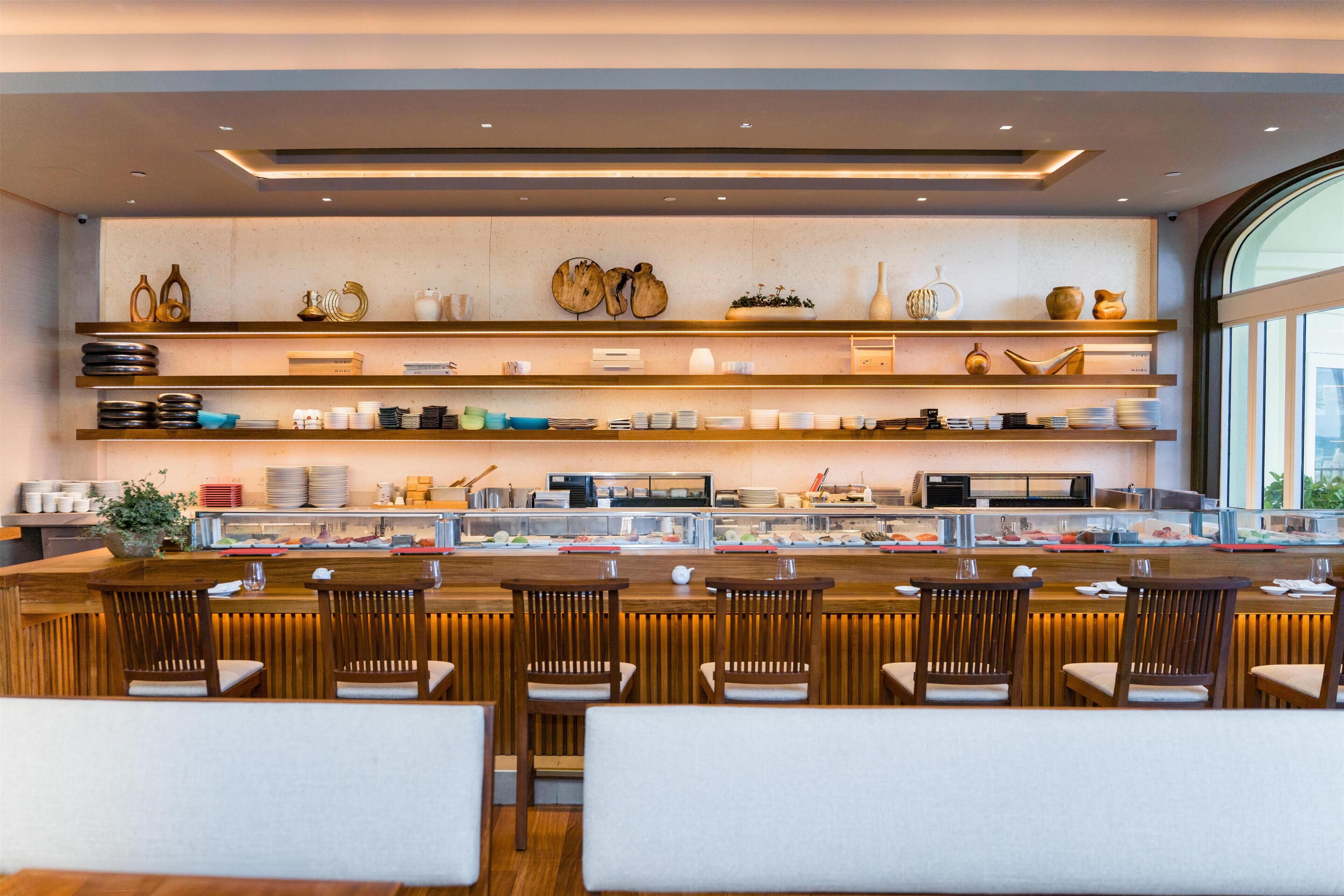 A modern restaurant with wooden bar and seating, shelves with dishes and decorative items, and a large window.