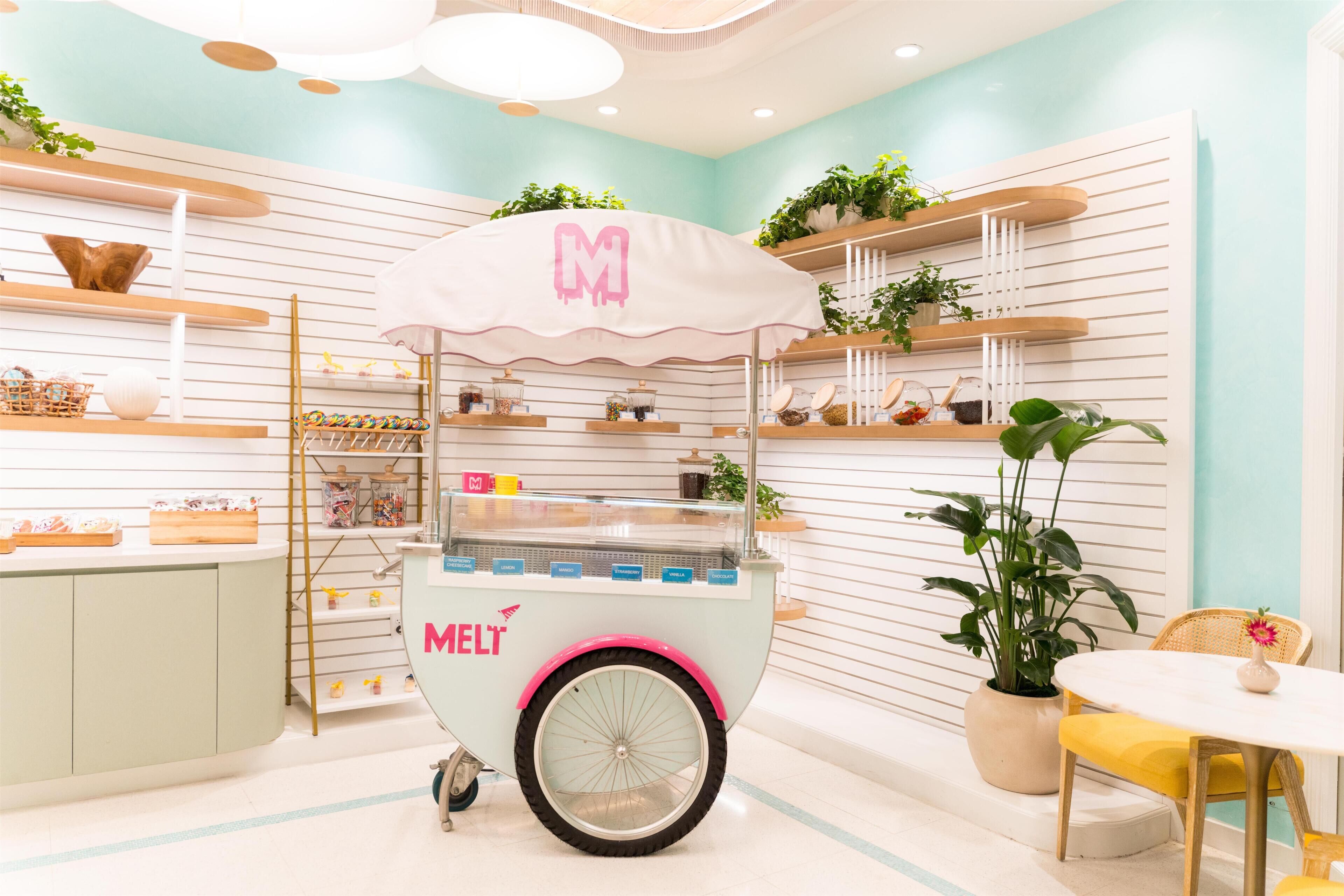 A mint green kiosk with a pink awning labeled 'MELT' in a brightly lit indoor space with white walls and shelves displaying various items.