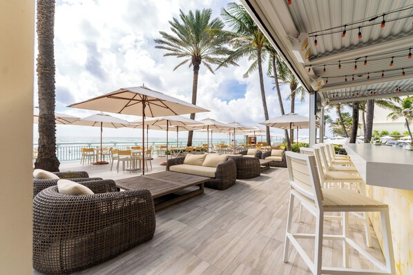 A beachside lounge with wicker sofas, white umbrellas, and a bar area. - Eau Resort & Spa (Manalapan)