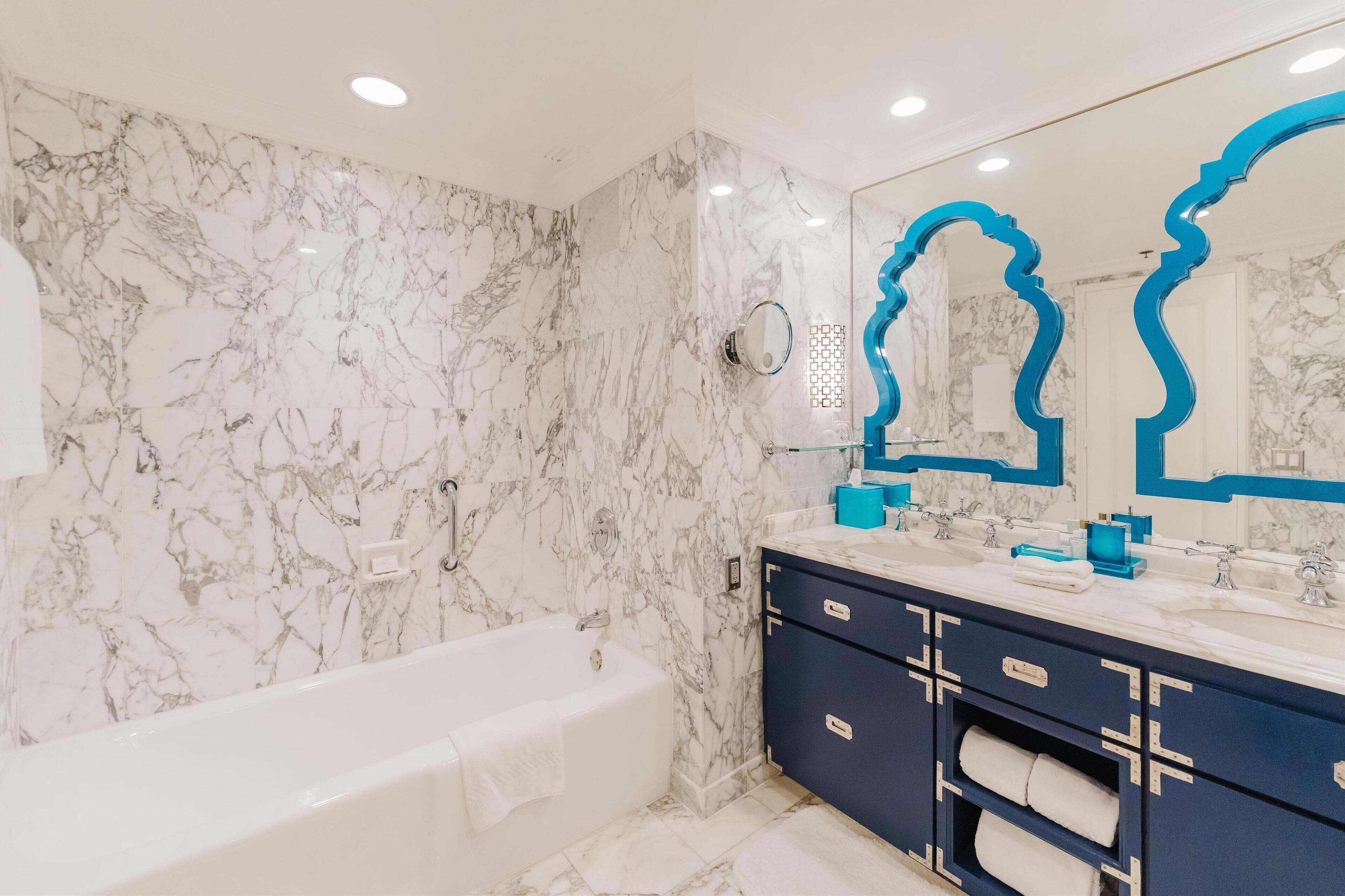 Un baño con pared de mármol, una bañera, un tocador azul con dos lavamanos y un espejo grande con marco azul.