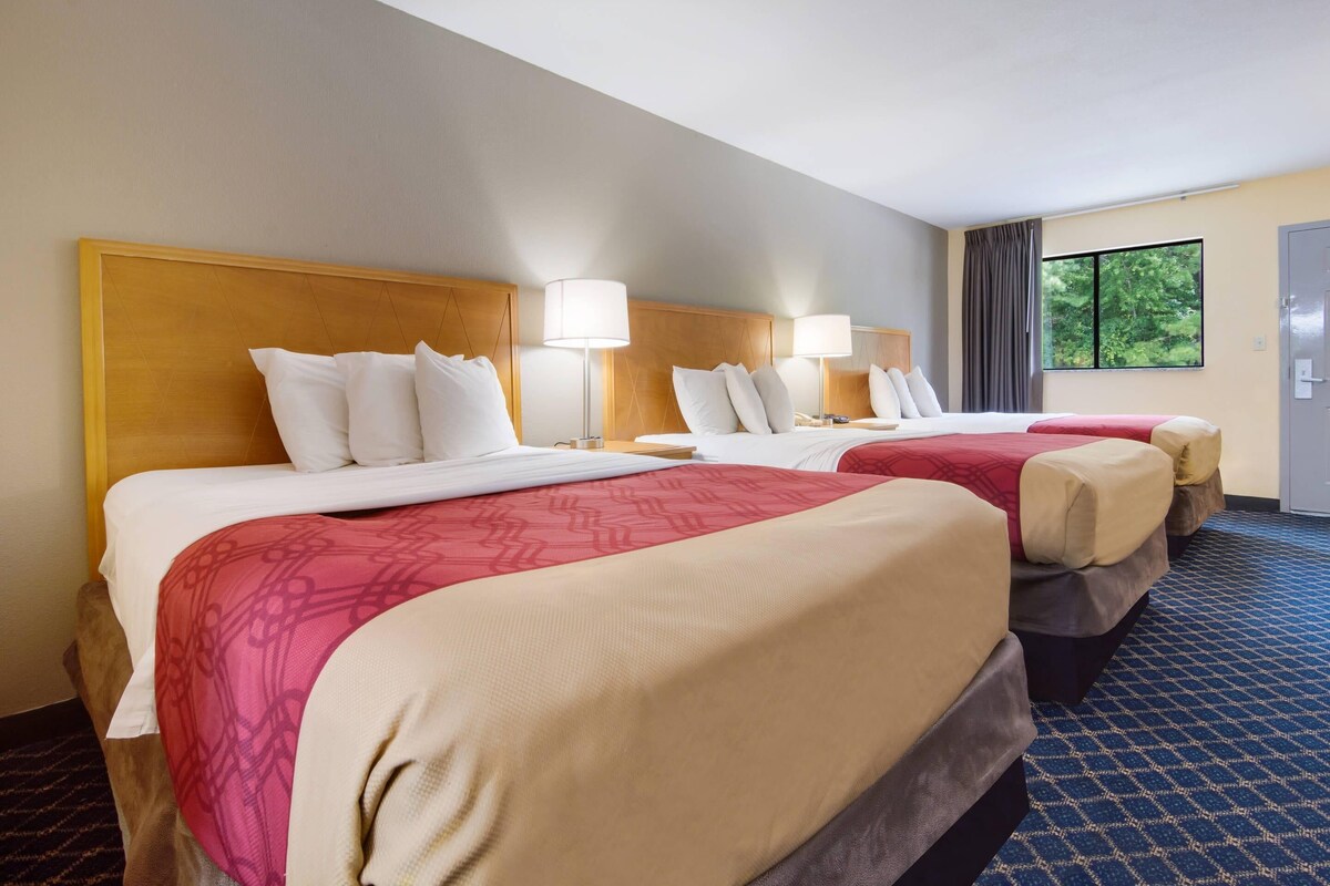 A hotel room with two beds, each with a wooden headboard, white bedding, and a red accent. There are bedside lamps and a window with a view of trees.