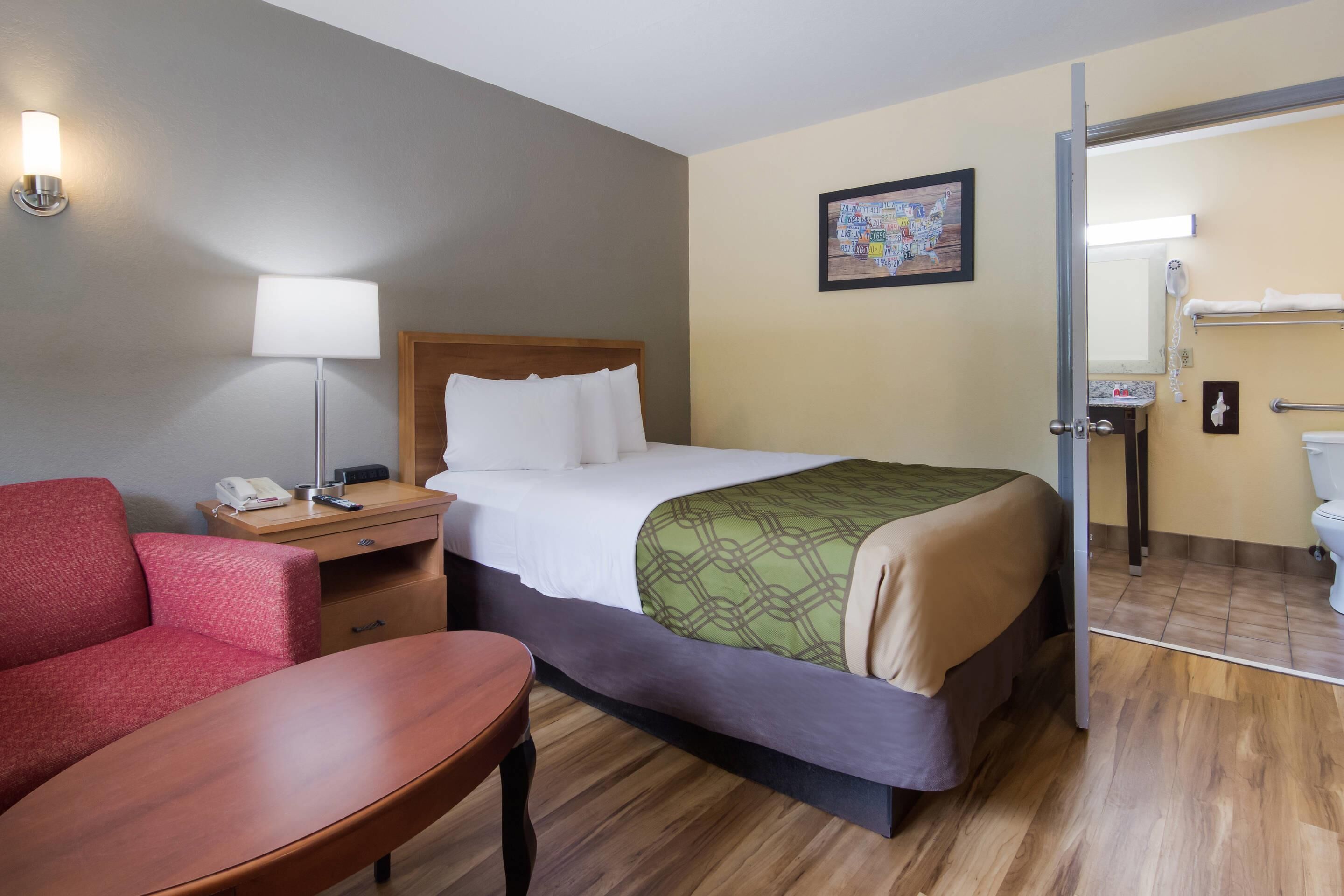 A hotel room with a bed, a desk, a chair, a lamp, a painting, and a bathroom visible through an open door.