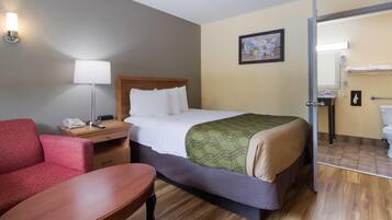A hotel room with a bed, a desk, a chair, a lamp, a painting, and a bathroom visible through an open door.