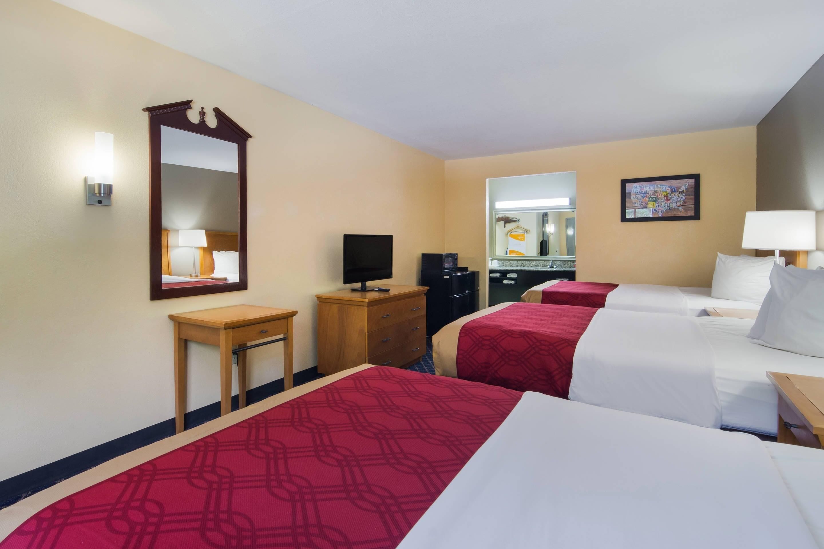 Habitación de hotel con dos camas, un escritorio, un espejo, televisión y baño visible a través de una puerta abierta.