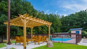 Una pérgola de madera con asientos, una piscina y un área cercada.