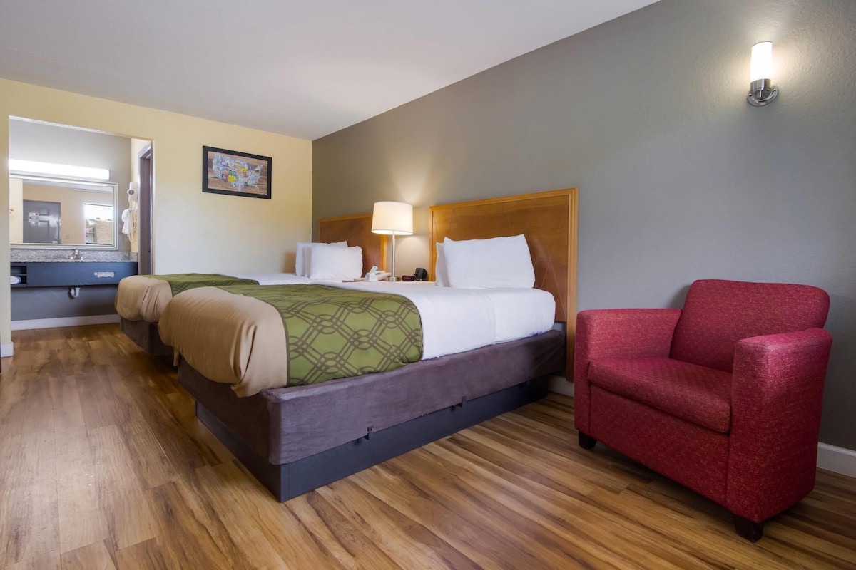 A hotel room with a bed, a red armchair, a wooden floor, a bathroom with a mirror, and a wall-mounted light fixture.
